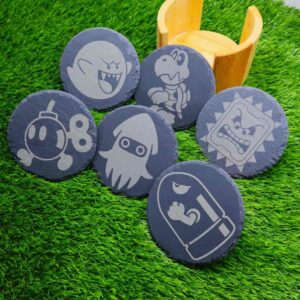 Bases de porta-copos de ardósia com inimigos icônicos de Super Mario, incluindo Thwomp, Bob-omb, Bullet Bill, Dry Bones, Boo e Blooper Squid, armazenadas em uma elegante caixa de bambu. Slate coasters featuring iconic Super Mario enemies, like Thwomp, Bob-omb, Bullet Bill, Dry Bones, Boo, and Blooper Squid, stored in a stylish bamboo case.
